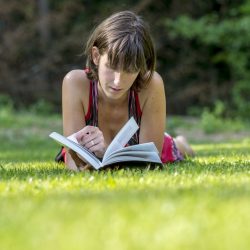 Young woman in summer dress lying in a green grass reading a book or novel.